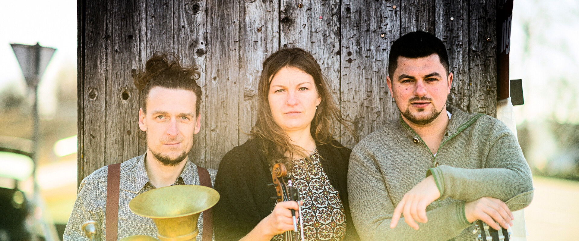 Drei Musiker posieren vor einer rustikalen Holzwand. Einer hält eine Tuba, eine andere eine Violine, und der dritte lehnt mit einer Gitarre. Das Sonnenlicht erzeugt einen warmen, natürlichen Effekt. | © Christoph Eder